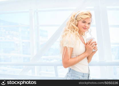 Beautiful young woman with music headphones, standing on the bridge with a take away coffee cup and posing against urban background.