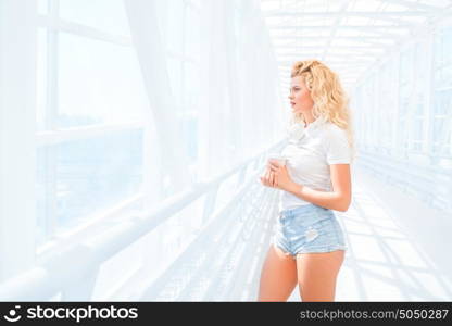 Beautiful young woman with music headphones, standing on the bridge with a take away coffee cup and posing against urban background.