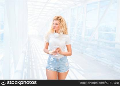 Beautiful young woman with music headphones, standing on the bridge with a take away coffee cup and posing against urban background.