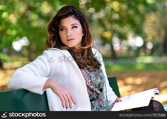Beautiful young woman with nice hairstyle sitting on a bench in the park and reading a book on a sunny autumn day.