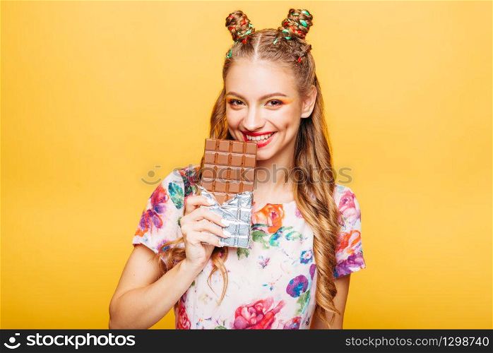 Beautiful young woman with playful look bites huge chocolate. Stylish girl with blonde curly hair. Stylish girl in colorful summer dress, yellow wall on background.. Woman with playful look bites huge chocolate