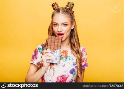 Beautiful young woman with playful look bites huge chocolate. Stylish girl with blonde curly hair. Stylish girl in colorful summer dress, yellow wall on background.. Woman with playful look bites huge chocolate