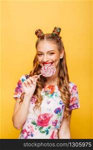 Beautiful young woman with playful look eating candy and smiling. Stylish girl with blonde curly hair. Portrait of attractive lady with big lollypop, yellow wall on background.. Woman with playful look eating candy