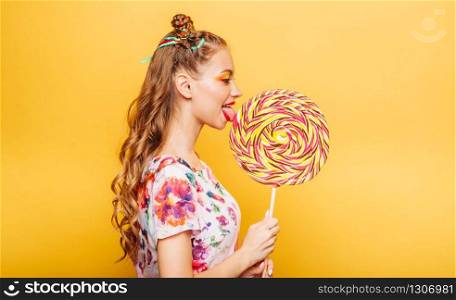 Beautiful young woman with playful look eating huge candy and smiling. Stylish girl with blonde curly hair. Portrait of attractive lady with big lollypop, yellow wall on background.. Woman with playful look eating candy