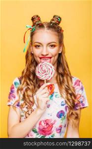 Beautiful young woman with playful look lick candy with tongue. Stylish girl with blonde curly hair. Portrait of attractive lady with big lollypop, yellow wall on background.. Woman with playful look lick candy with tongue