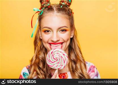 Beautiful young woman with playful look lick candy with tongue. Stylish girl with blonde curly hair. Portrait of attractive lady with big lollypop, yellow wall on background.. Woman with playful look lick candy with tongue
