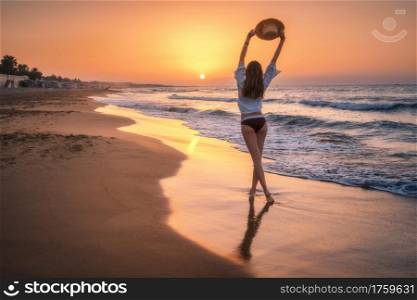 Beautiful young woman with raised up arms with straw hat on sandy beach at colorful sunset. Sea with waves. Summer travel and resort. Happy slim girl in bikini on the tropical sea coast. Lifestyle