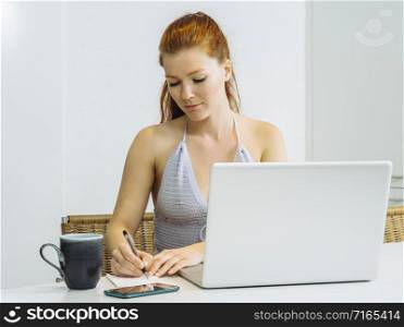 Beautiful young woman with red hair sitting with a laptop and writing on a notepad.