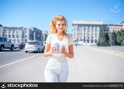 Beautiful young woman with two takeaway coffee cups, standing against urban city background and looking aside.