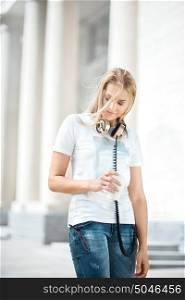 Beautiful young woman with vintage music headphones around her neck, holding a take away coffee and standing with wind in her hair against urban city background.