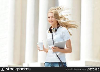 Beautiful young woman with vintage music headphones around her neck, holding a take away coffee and standing with wind in her hair against urban city background.