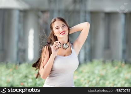 Beautiful young woman with vintage music headphones around her neck, posing against nature background and smiling happily.