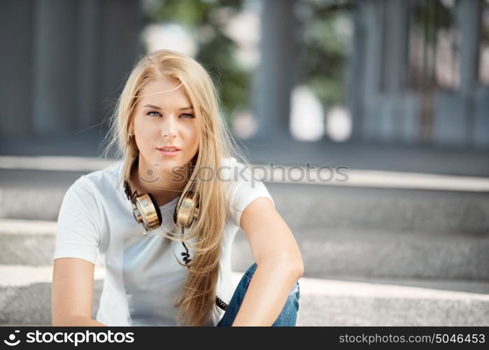 Beautiful young woman with vintage music headphones around her neck, sitting against urban city background and looking straight.