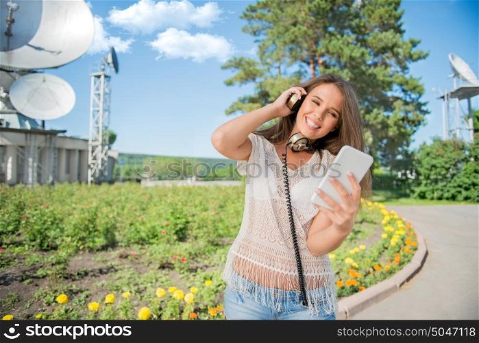 Beautiful young woman with vintage music headphones around her neck, surfing internet on a smartphone and standing against background of satellite dishes that receives wireless signals from satellites.