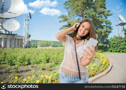 Beautiful young woman with vintage music headphones around her neck, surfing internet on a smartphone and standing against background of satellite dishes that receives wireless signals from satellites.