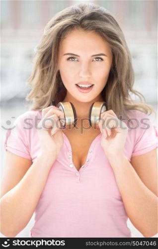Beautiful young woman with vintage music headphones on her neck, standing against urban city background and listening to the music.