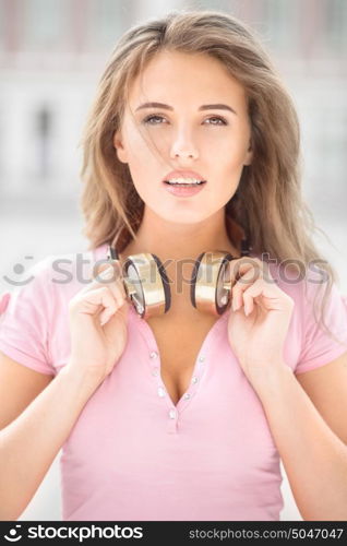 Beautiful young woman with vintage music headphones on her neck, standing against urban city background and listening to the music.