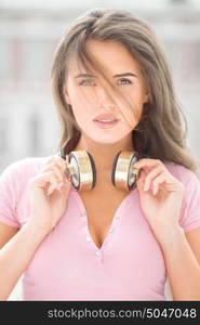 Beautiful young woman with vintage music headphones on her neck, standing against urban city background and listening to the music.