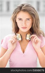 Beautiful young woman with vintage music headphones on her neck, standing against urban city background and listening to the music.
