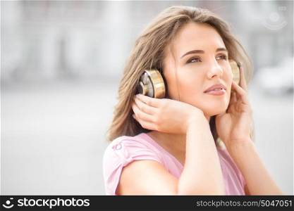 Beautiful young woman with vintage music headphones, standing against urban city background and listening to the music.