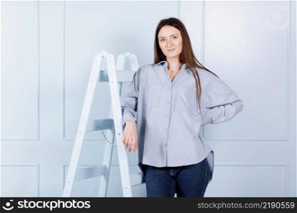 beautiful young woman with with long hair in blue clothes rests on a stepladder on blue wall background. beautiful young woman with with long hair in blue clothes rests on a stepladder on blue wall background.