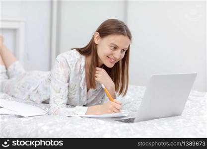 beautiful Young Woman Working from Home - Female Entrepreneur on Bed with Laptop Computer, Paperwork from Home. happy girl looks at laptop and makes notes. quarantine. coronavirus. beautiful Young Woman Working from Home - Female Entrepreneur on Bed with Laptop Computer, Paperwork from Home. happy girl looks at laptop and makes notes. quarantine. coronavirus.