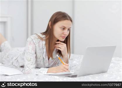 Beautiful young woman working, using laptop computer and makes notes in the bedroom at home. Freelancer. Writing, typing. Girl checking social apps. Communication and technology concept.. Beautiful young woman working, using laptop computer and makes notes in the bedroom at home. Freelancer. Writing, typing. Girl checking social apps. Communication and technology concept