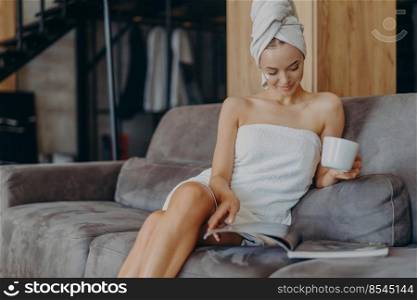 Beautiful young woman wrapped in bath towel, reads beauty magazine, drinks hot beverage, poses on comfortable couch in living room, applies face cream, has healthy smooth skin after taking shower