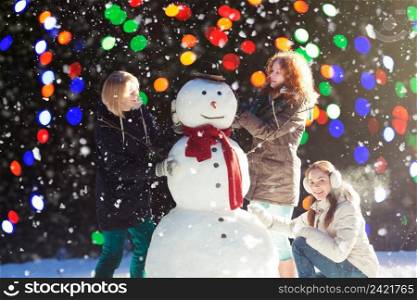 Beautiful young women enjoying building a snowman on a snowy winter day, blurred lights on background