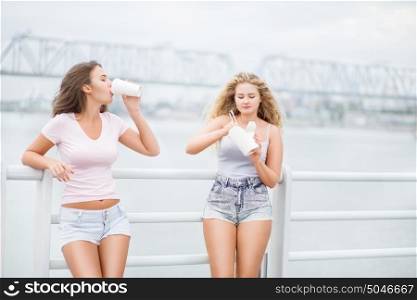 Beautiful young women, leaning on parapet, sharing a fast food lunch box, eating up noodles from Chinese take-out with chopsticks and drinking takeaway coffee.