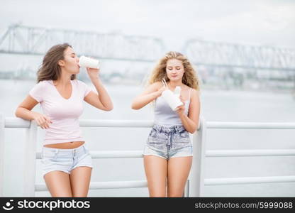 Beautiful young women, leaning on parapet, sharing a fast food lunch box, eating up noodles from Chinese take-out with chopsticks and drinking takeaway coffee.