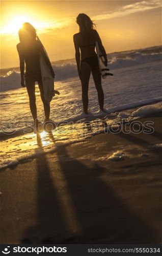 Beautiful young women surfer girls in bikinis with surfboards on a beach at sunset or sunrise