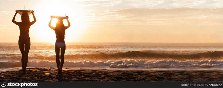 Beautiful young women surfer girls in bikinis with surfboards on a beach at sunset or sunrise panoramic web banner