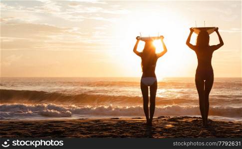 Beautiful young women surfer girls in bikinis with surfboards on a beach at sunset or sunrise