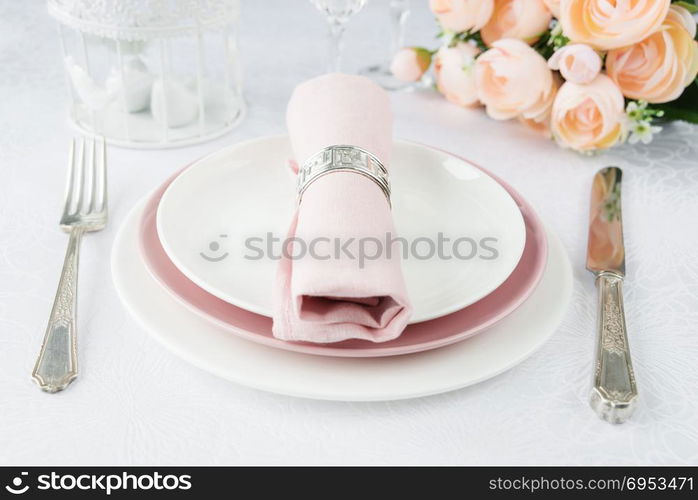 Beautifully decorated table with white and pink plates, glasses, silverware, pink napkin and flowers on luxurious tablecloths