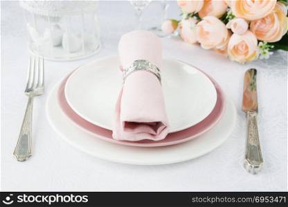 Beautifully decorated table with white and pink plates, glasses, silverware, pink napkin and flowers on luxurious tablecloths