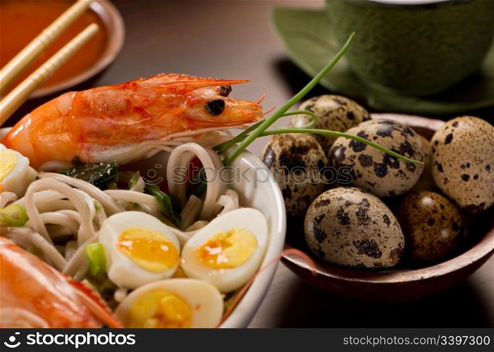 Beautifully prepared prawns and quail eggs served over noodles. Chopsticks, a bowl with sauce, a tea cup and a bowl of quail eggs are also on the table.