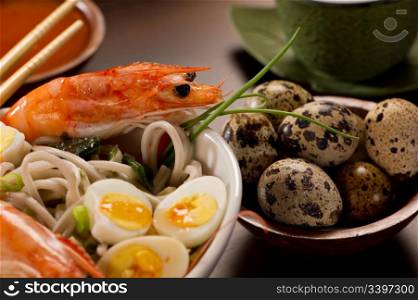 Beautifully prepared prawns and quail eggs served over noodles. Chopsticks, a bowl with sauce, a tea cup and a bowl of quail eggs are also on the table.
