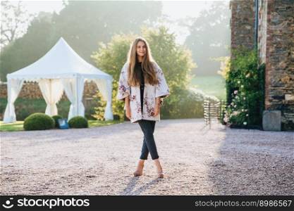 Beautisful stylish woman wears fashionable clothes, has long straight hair, stand crossed legs against beautiful nature, ancient builduing and white tent enjoys sunshine has excursion in unknown place