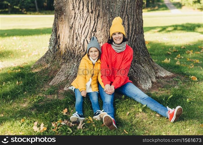 Beautiul female wears knitted hat and swetaer sits together with her little daughter near tree, have rest outdoors in beautiful park, enjoy fresh autumn air, feel relaxed. People, relaxation