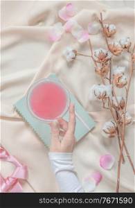 Beauty aesthetic lifestyle with women hand holding cocktail glass on beige fabric background with cotton branches and pink petals. Top view