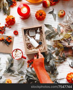 Beauty and autumn skin care concept. Female hand in orange knitted sweater opens beauty box with cosmetic products on desktop with hot drink, fall leaves, pumpkins and burning candles. Top view