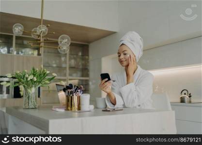 Beauty and facial care concept. Young pretty woman in white bathrobe applying cosmetic product on face and looking in compact mirror, taking care of her skin while sitting in modern kitchen at home. Happy healthy woman in bathrobe doing skincare daily routine after taking shower or bath at home