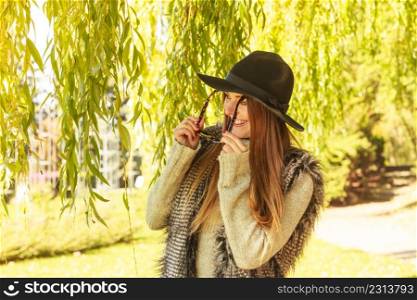Beauty and fashion of women. Young attractive fashionable girl wearing stylish hat waistcoat and sunglasses. Pretty woman around leaves of willow tree.. Adorable woman in sunglasses
