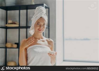 Beauty and hygiene concept. Attractive healthy young woman has shiny perfect smooth skin, applies body cream, holds jar of cosmetic product, stands wrapped in bath towel, poses in cozy bathroom
