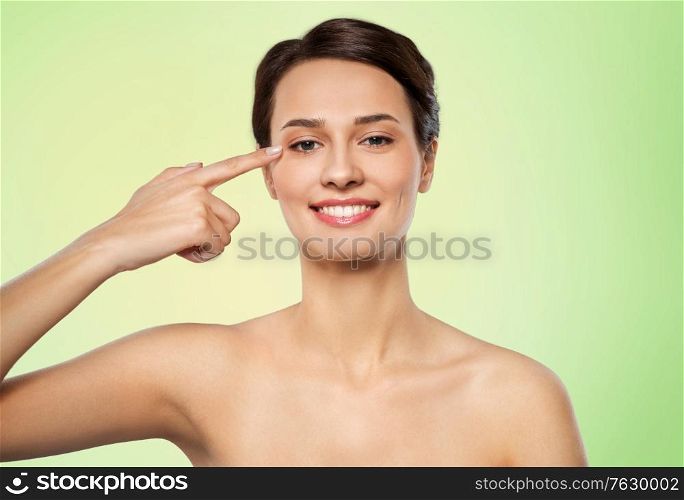 beauty and people concept - beautiful young woman with bare shoulders pointing to her eye over lime green natural background. beautiful young woman pointing to her eye