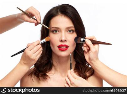 beauty and people concept - beautiful young woman with red lipstick and hands of make up artists with brushes over white background. beautiful smiling young woman with red lipstick