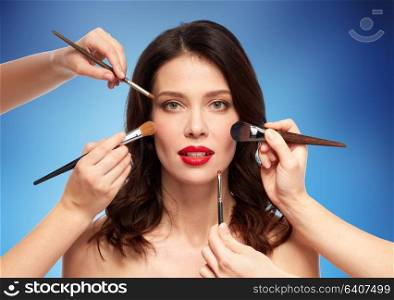 beauty and people concept - beautiful young woman with red lipstick and hands of make up artists with brushes over blue background. woman and hands of make up artists with brushes