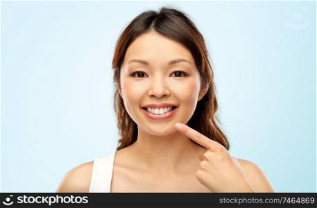 beauty and people concept - happy smiling young asian woman pointing to her mouth or teeth over blue background. happy smiling young asian woman touching her face