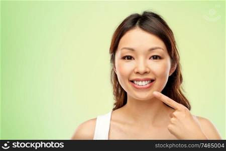 beauty and people concept - happy smiling young asian woman pointing to her mouth or teeth over lime green natural background. happy smiling young asian woman touching her face